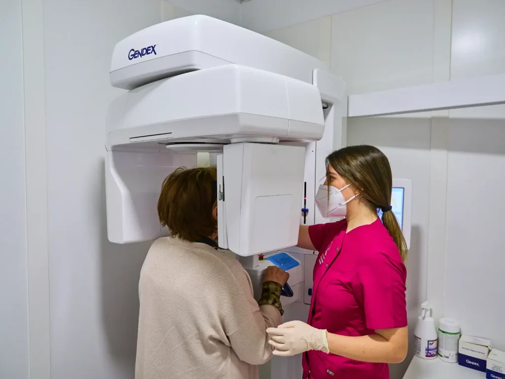 Técnico dental con uniforme fucsia y mascarilla asistiendo a una paciente para realizarle una radiografía panorámica u ortopantomografía con una máquina Gendex.
