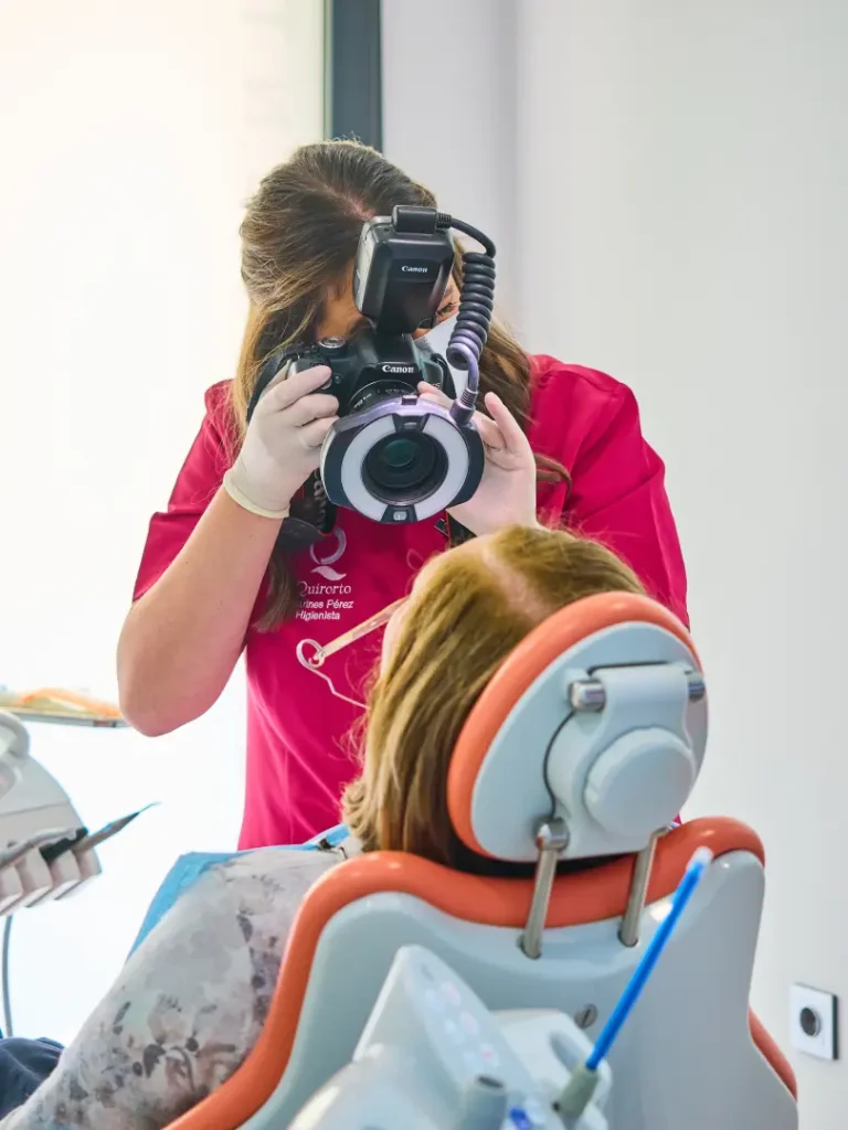 Técnico dental con uniforme fucsia tomando una fotografía clínica intraoral de una paciente en el sillón dental utilizando una cámara DSLR con flash anular profesional.