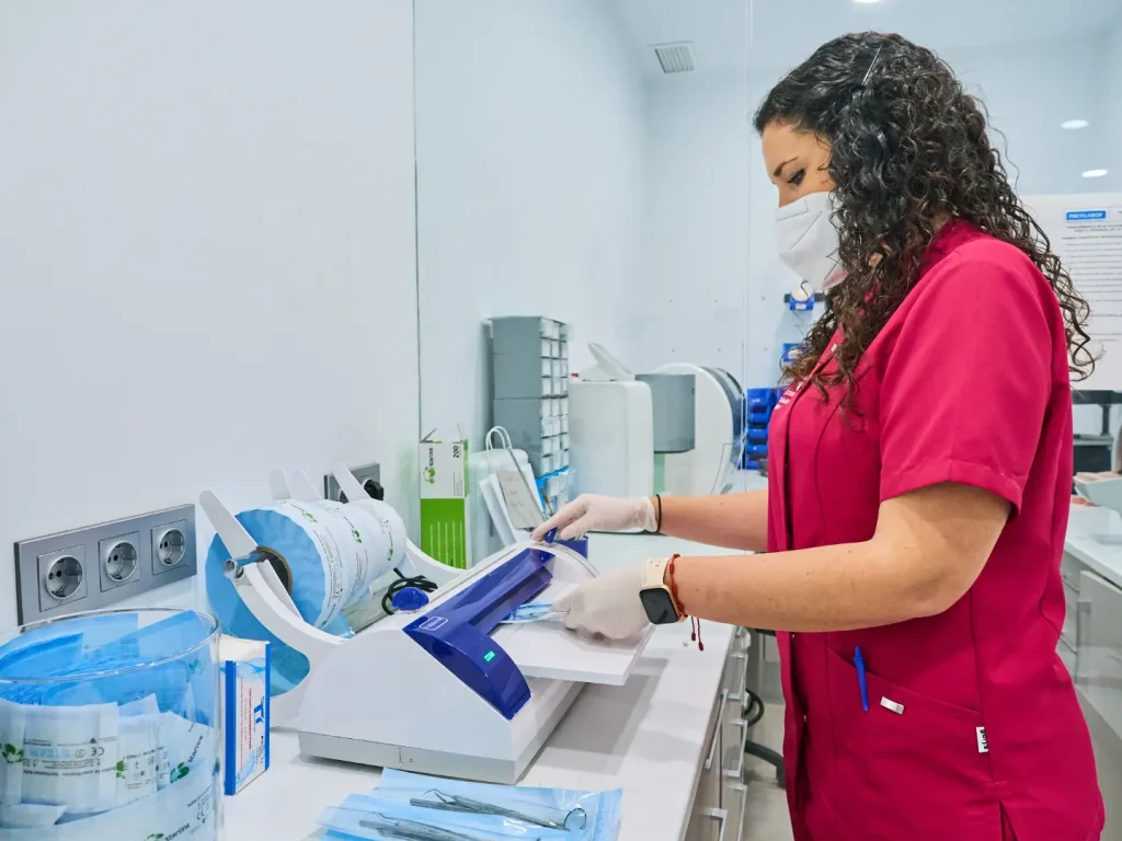 Técnico dental con uniforme fucsia y mascarilla sellando paquetes de esterilización de instrumental con una máquina termoselladora en la clínica.