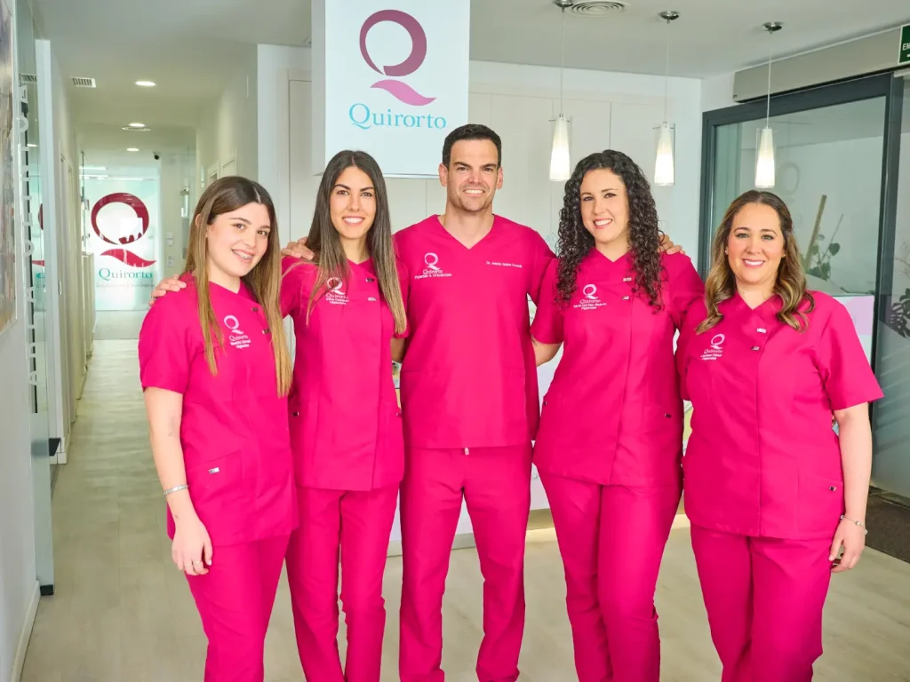 Equipo médico de la clínica dental Quirorto, compuesto por cinco profesionales sonrientes, vestidos con uniformes fucsia brillante, posando en el área de recepción con el logo de la clínica visible detrás.