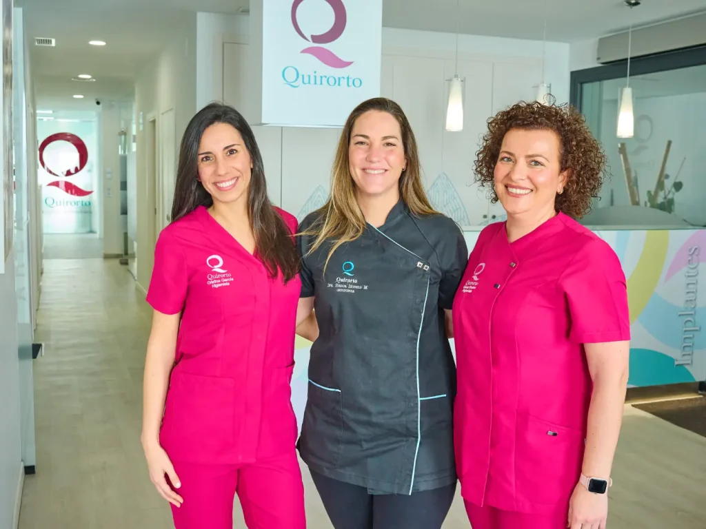 Tres mujeres del equipo de la clínica Quirorto, dos con uniforme fucsia y una en el centro con uniforme negro y gris, posando sonrientes frente a la recepción y el logo de la clínica.