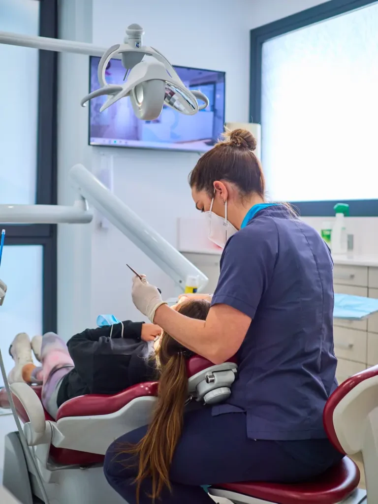 Dentista o higienista con uniforme azul oscuro y mascarilla, tratando a una paciente recostada en el sillón dental, bajo la luz de trabajo y con un monitor mostrando imágenes clínicas de fondo.