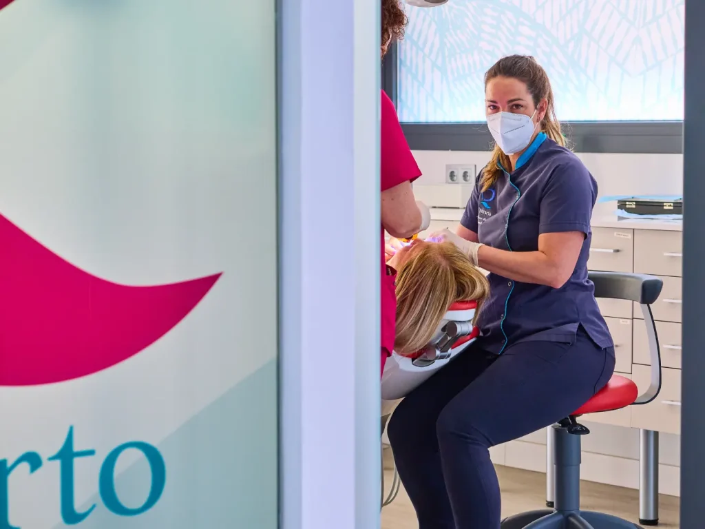Dentista con uniforme azul oscuro y mascarilla, tratando a una paciente en el sillón dental, mientras otra asistente con uniforme fucsia ayuda, con un gran detalle del logo de la clínica en primer plano.