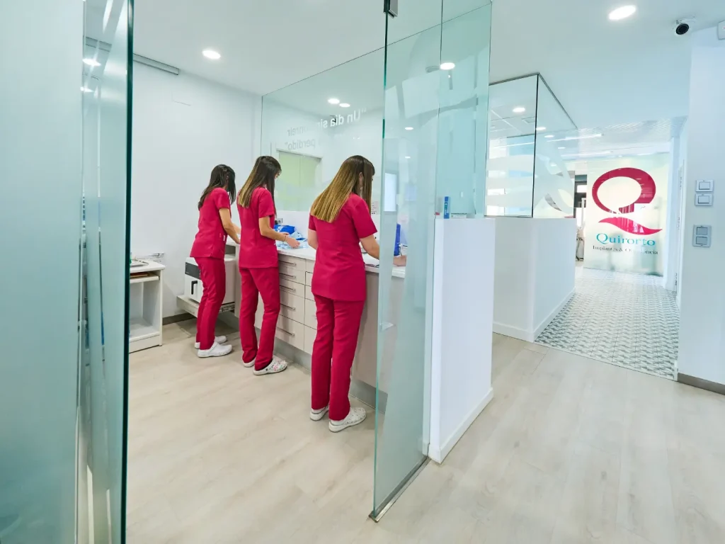 Tres miembros del equipo de la clínica Quirorto, vestidas con uniformes fucsia, trabajando en una estación de apoyo u oficina, separadas por paneles de vidrio, con el logo de la clínica visible al fondo.