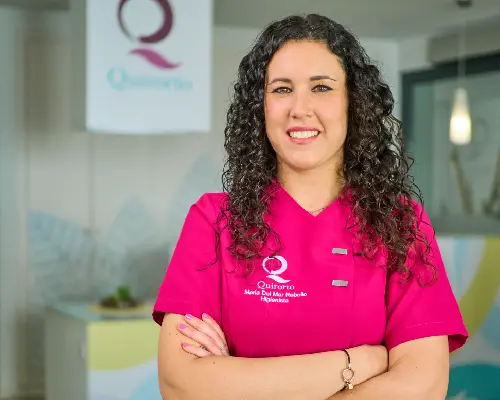 Retrato de primer plano de una odontóloga con cabello rizado y uniforme médico rosa fuerte con el logo de "Quirorto", sonriendo con los brazos cruzados en un ambiente de clínica moderna.