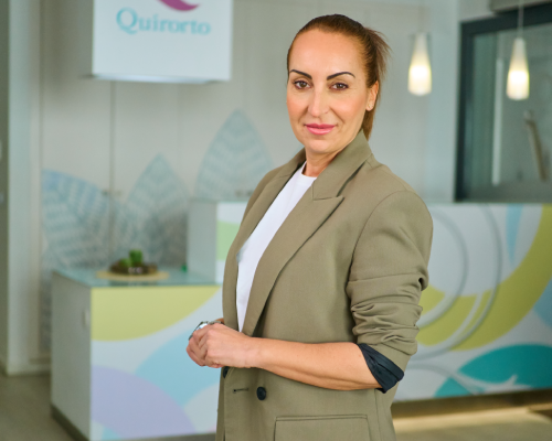 Retrato de plano medio de una mujer con chaqueta color caqui y cabello recogido, posando seria pero amable en la recepción de la clínica Quirorto con el mostrador colorido detrás.