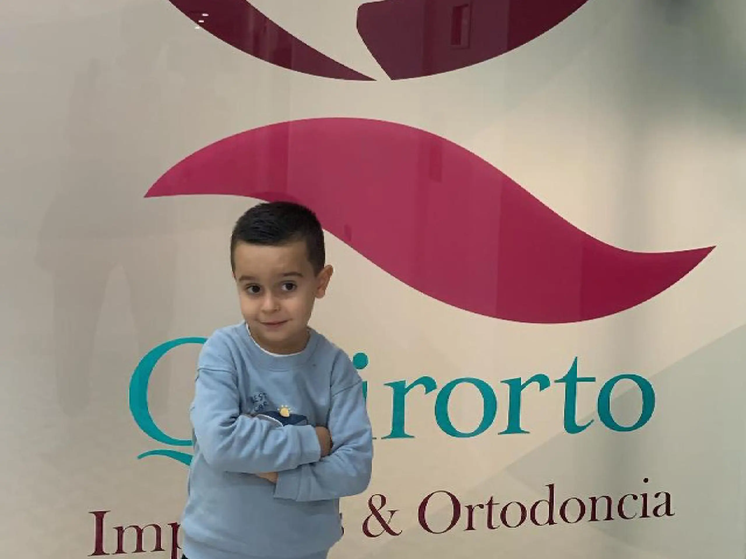 Niño pequeño con los brazos cruzados posando frente a un logotipo de clínica dental que incluye las palabras "Qirorto" e "Implantes & Ortodoncia".