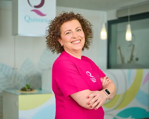 Mujer de mediana edad con cabello rizado y castaño, vestida con una bata médica rosa fuerte con el logo "Quirorto", sonriendo con los brazos cruzados en un entorno de clínica.