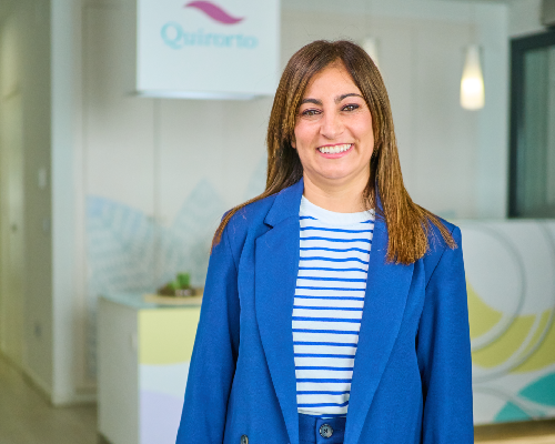 Retrato de una mujer sonriente con chaqueta azul eléctrico y camiseta a rayas blancas y azules, posando en la recepción de la clínica Quirorto con su logo de fondo.