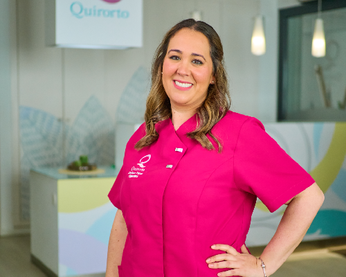 Retrato de plano medio de una profesional dental sonriente de la clínica Quirorto, vestida con el uniforme fucsia, con el mostrador de recepción de colores pastel detrás.
