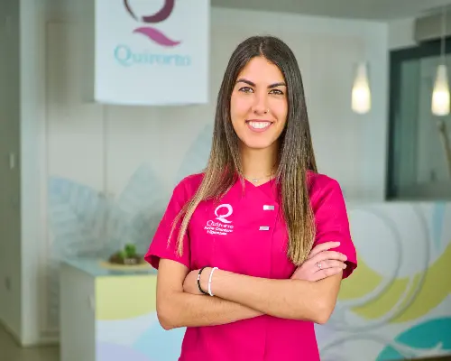 Retrato de mujer joven con cabello largo y oscuro y uniforme médico rosa fuerte de Quirorto, sonriendo con los brazos cruzados en la recepción de la clínica.