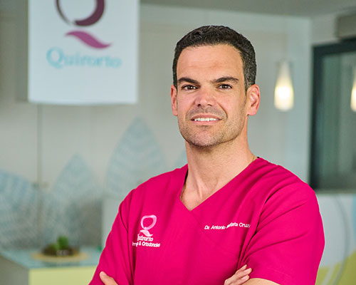 Retrato de plano medio de un dentista, el Dr. Antonio Batista, con uniforme sanitario fucsia y brazos cruzados, posando sonriente frente al logo de la clínica Quirorto.