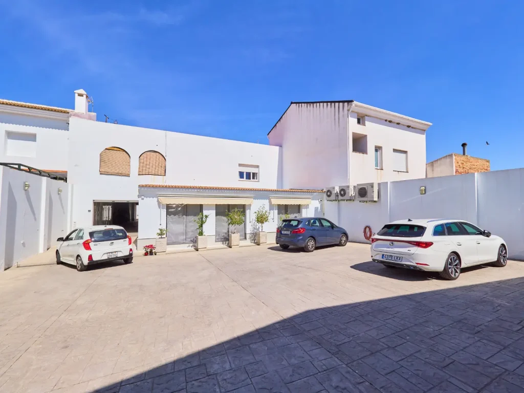 Vista exterior de un edificio blanco, probablemente la clínica, con una explanada de parking con tres coches estacionados, bajo un cielo azul brillante.