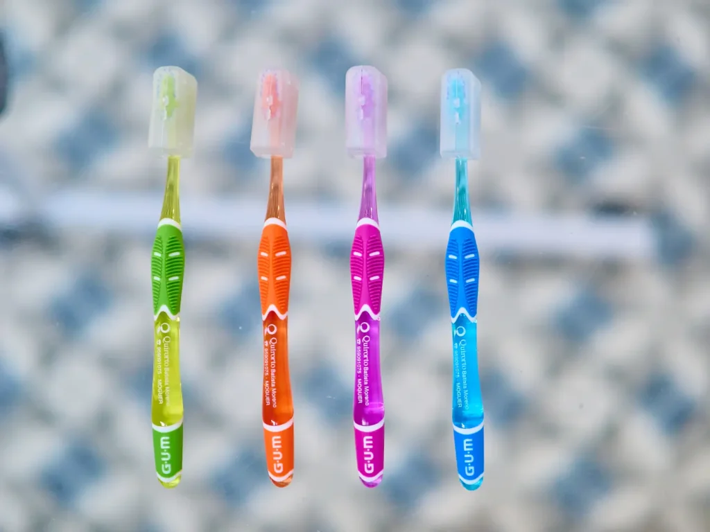 Cuatro cepillos de dientes de la marca GUM, de colores neón (verde, naranja, morado y azul), con protectores de cabeza de plástico transparente, sobre una superficie.