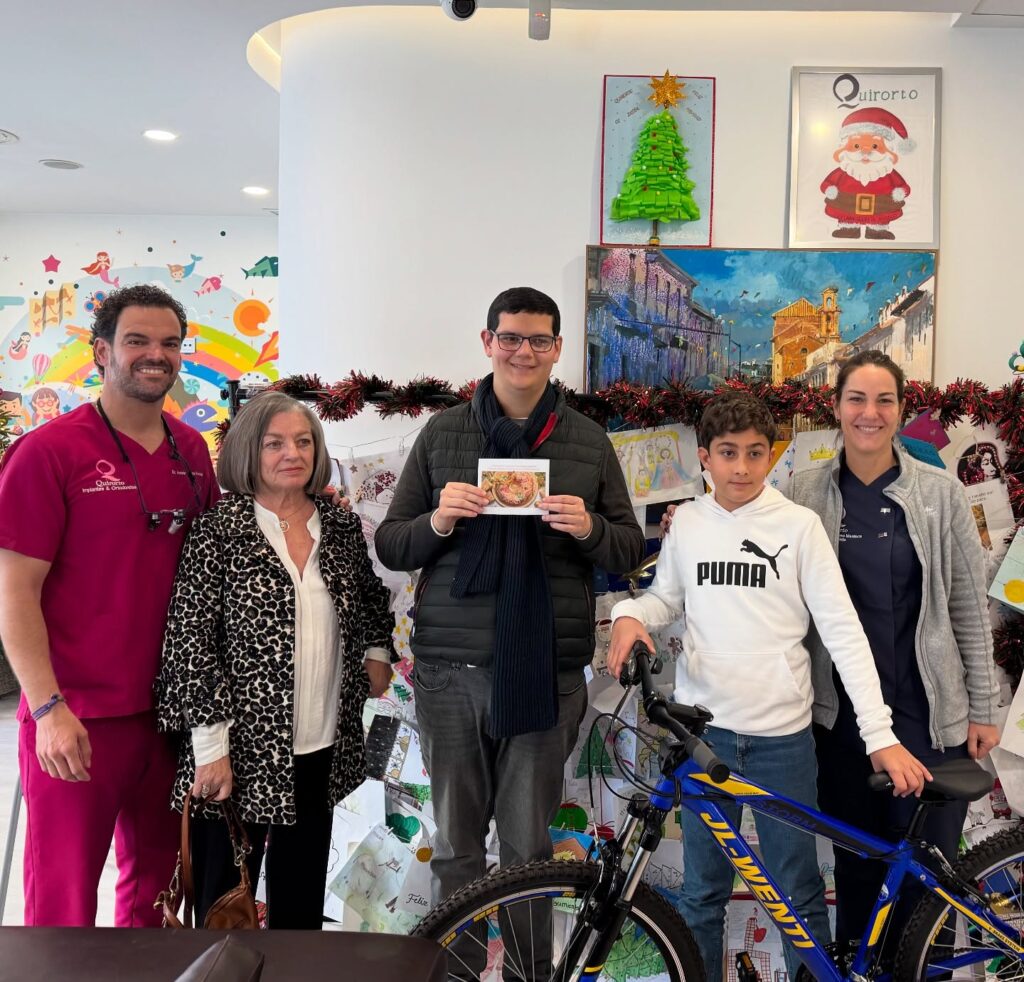 Cinco personas (dos miembros del personal de Quiroorto y tres pacientes/familiares, incluyendo un niño con una bicicleta) posando en la clínica durante la temporada navideña, con un árbol y un cartel de Santa Claus.