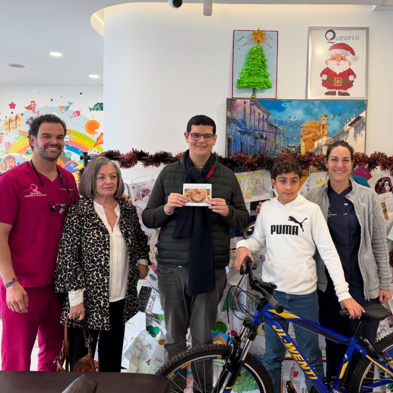 Cinco personas (dos miembros del personal de Quiroorto y tres pacientes/familiares, incluyendo un niño con una bicicleta) posando en la clínica durante la temporada navideña, con un árbol y un cartel de Santa Claus.