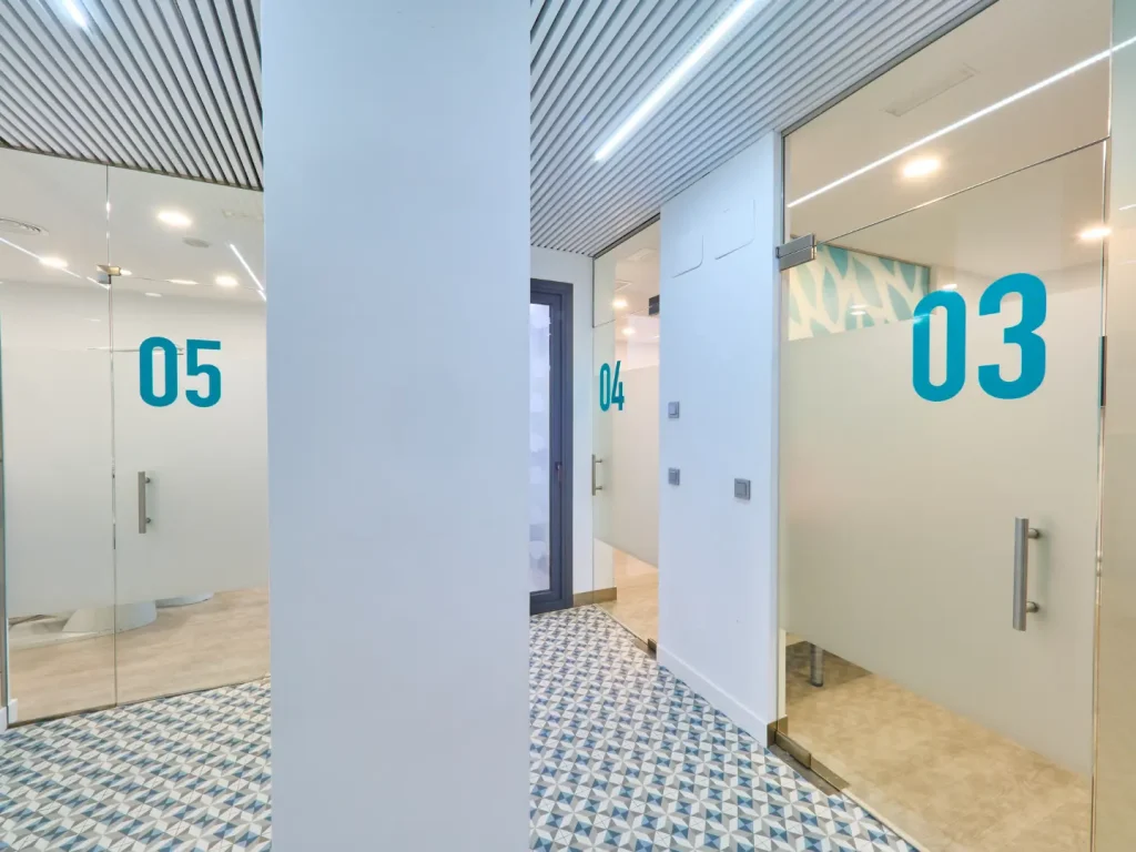 Pasillo moderno con suelo de baldosas geométricas azules y blancas, y puertas de vidrio esmerilado numeradas con grandes dígitos turquesa (05, 04, 03) que conducen a las salas de tratamiento.