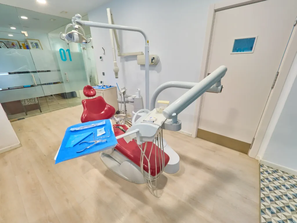 Gabinete dental número 01 con un sillón de tratamiento rojo y blanco, instrumental quirúrgico dispuesto sobre una bandeja y pared con puerta blanca y baldosas geométricas azules.