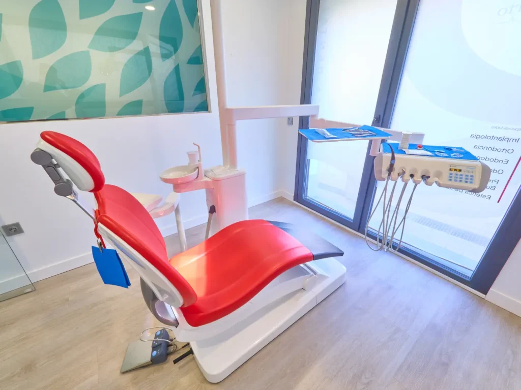 Sillón dental moderno de color rojo y blanco en un gabinete luminoso, con la unidad de instrumental, la escupidera y un gran ventanal de fondo.
