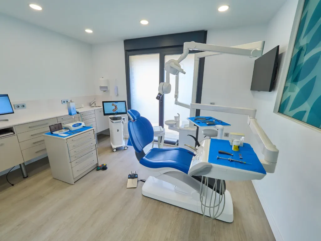 Gabinete dental moderno con sillón azul y blanco, equipamiento de alta tecnología, monitor para visualización de radiografías o escaneos y encimeras blancas con cajoneras.