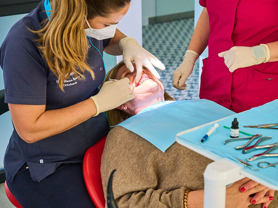 Odontóloga con uniforme azul y mascarilla trabajando en la boca de una paciente que lleva brackets, asistida por una auxiliar con uniforme rosa, en un consultorio dental.