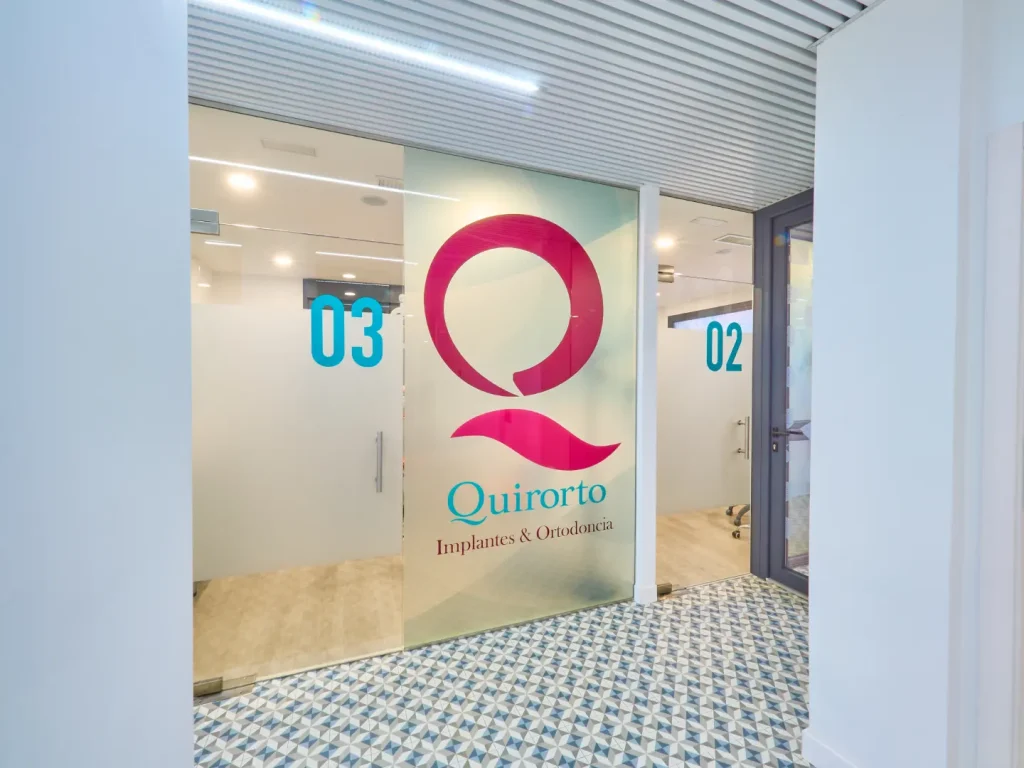 Puertas de vidrio esmerilado con el gran logo magenta de Quirorto y los números de gabinete en turquesa (03 y 02) en un pasillo con suelo de baldosas geométricas azules y blancas.