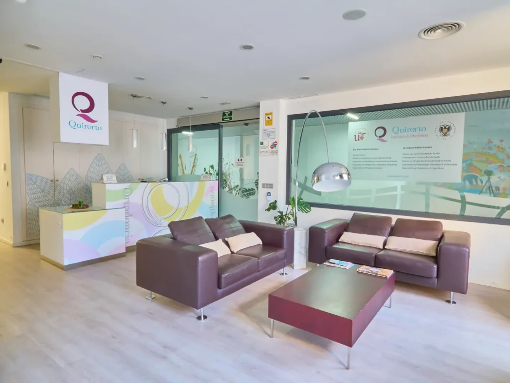 Sala de espera moderna y luminosa de la clínica dental Quirorto, con dos sofás de cuero marrón oscuro, una mesa de centro a juego, y el mostrador de recepción colorido al fondo.