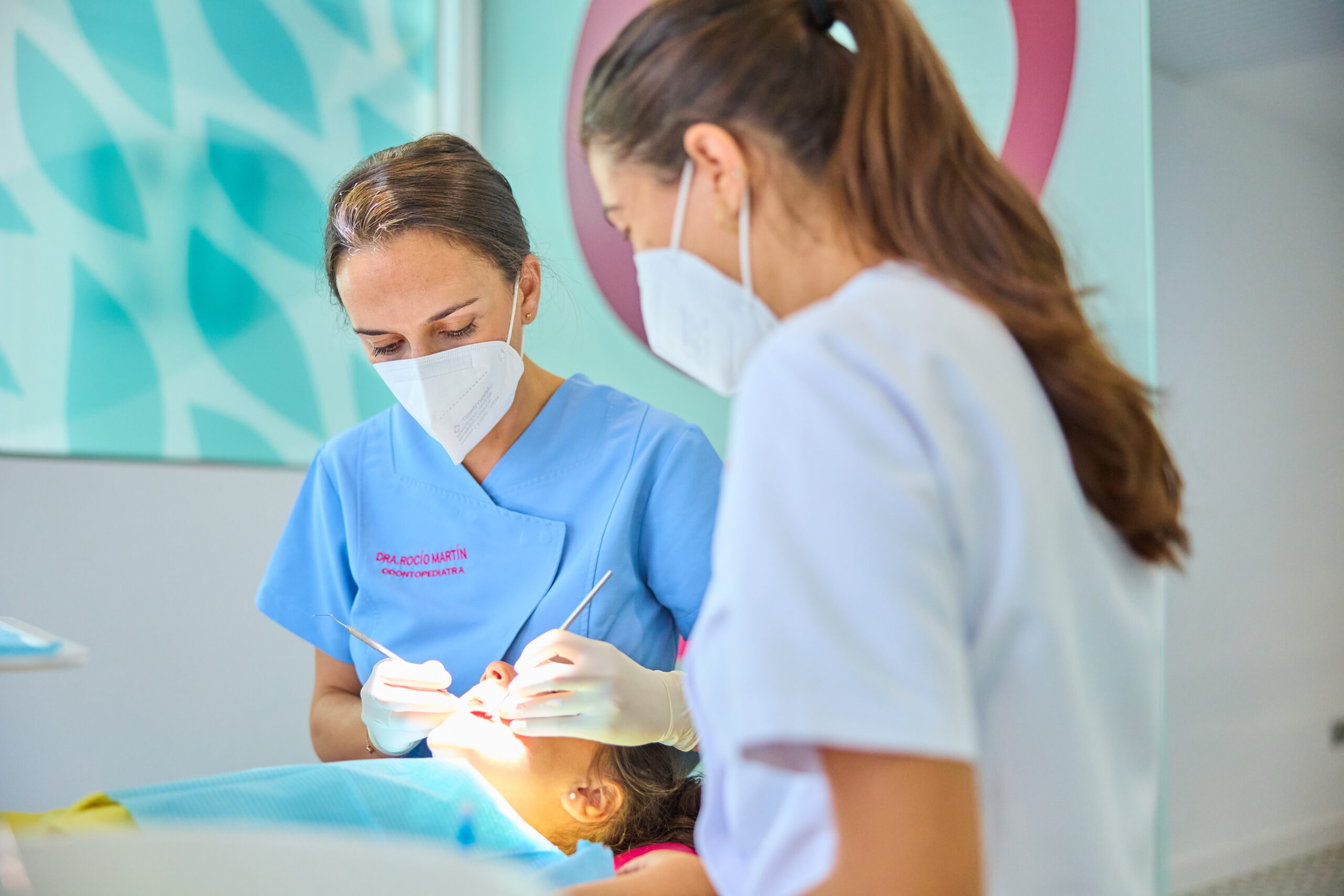 Dos dentistas o asistentes dentales, ambas con mascarillas y guantes, realizan un procedimiento dental a un paciente acostado, utilizando instrumentos. La doctora principal viste un uniforme azul claro.