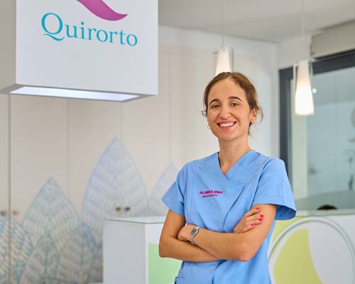 Retrato de plano medio de una dentista o profesional sanitaria con uniforme azul claro, sonriendo con los brazos cruzados, en el vestíbulo de la Clínica Quirorto.