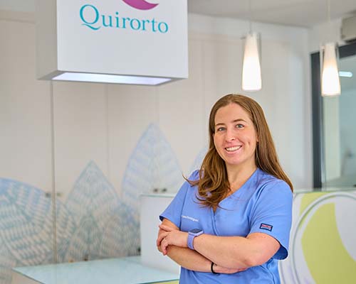 etrato de plano medio de una dentista o asistente dental con uniforme azul, sonriendo con los brazos cruzados, en el vestíbulo de la Clínica Quirorto.