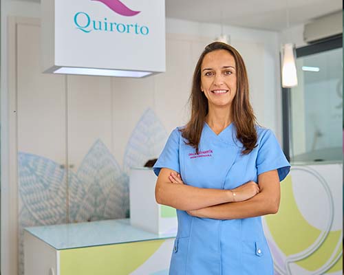 Retrato de plano medio de una dentista o profesional sanitaria con uniforme azul claro, sonriendo con los brazos cruzados, en el vestíbulo de la Clínica Quirorto.