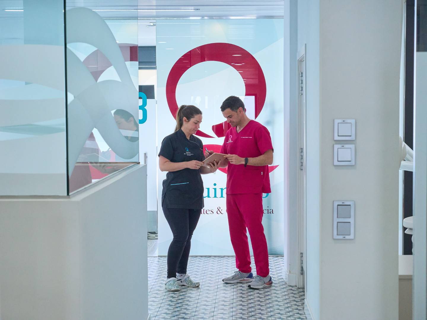 Dos profesionales de la salud, un hombre y una mujer con uniformes médicos, revisan un portapapeles o tableta en el pasillo de una clínica dental con paredes de vidrio y logotipos.